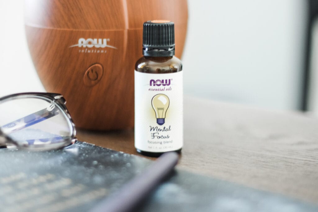 Image of essential oil blend mental focus. Bottle is on a desk with glasses and a diffuser in the background. 